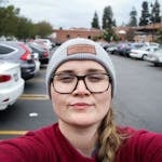 Portrait of a woman wearing glasses and a beanie, taking a selfie in a busy parking lot.